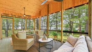 Une terrasse en bois avec des meubles en osier, un ventilateur de plafond et de grandes fenêtres donnant sur une zone boisée.