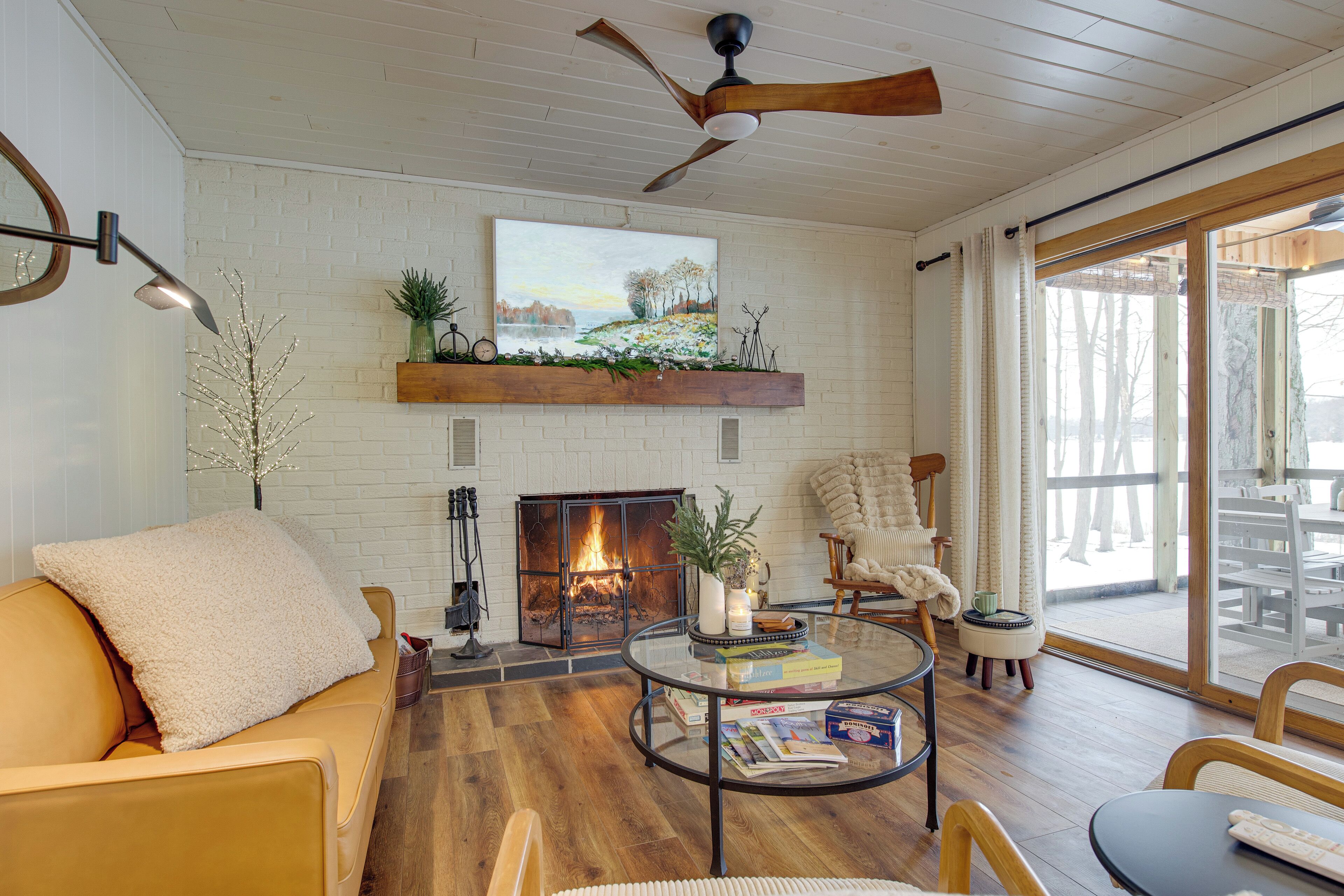 A cozy living room with a fireplace, wooden floors, a ceiling fan, and a large window.