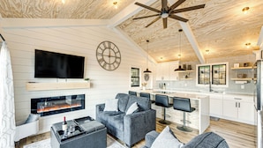 A modern living room with a fireplace, grey sofas, a flat-screen TV, and a kitchen area with white cabinets and a bar. - Canoes & Dock: Waterfront Retreat on Lake Beulah! (East Troy)