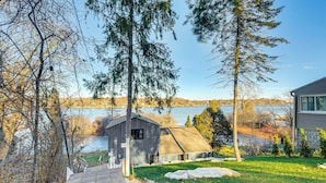 A wooden cabin with a sloped roof, a stone pathway leading to it, and a view of a lake and trees in the background. - Canoes & Dock: Waterfront Retreat on Lake Beulah! (East Troy)