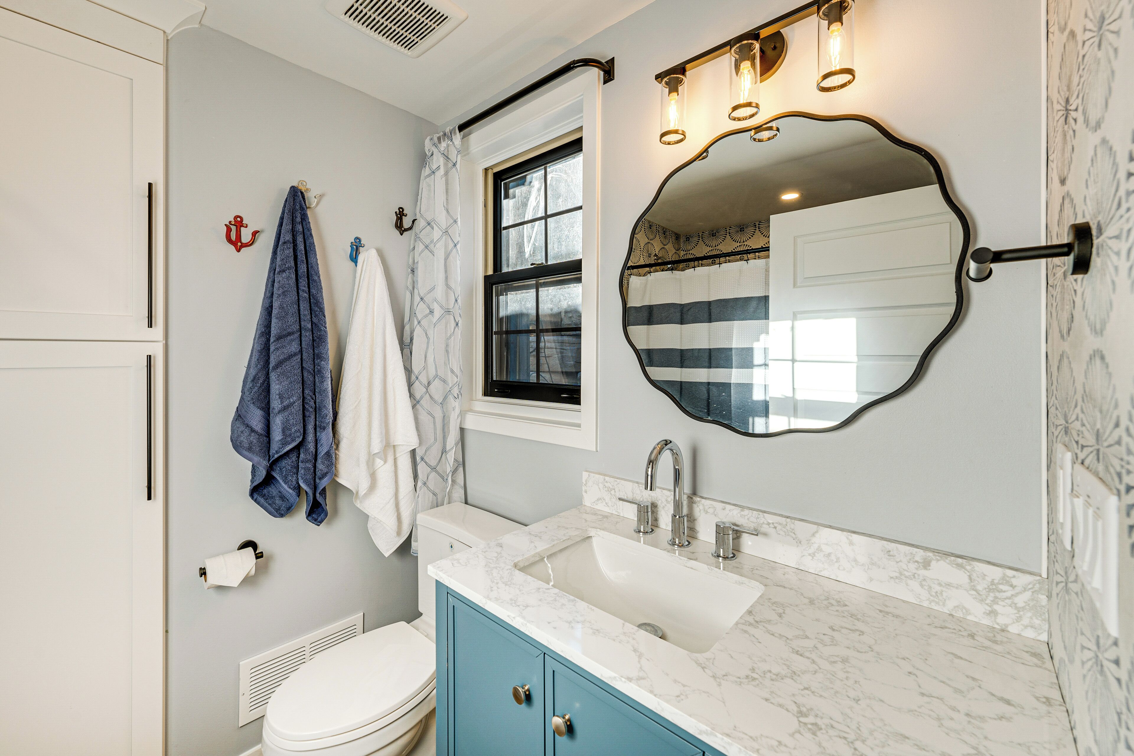 Un baño con inodoro blanco, un mueble azul, un lavabo con encimera de mármol, un espejo con luces y una ventana con cortina.