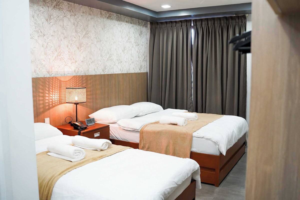 A hotel room with two beds, a nightstand with a lamp, and a large window with curtains.