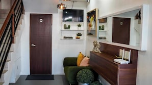 Un recibidor moderno con puerta de madera, una escalera, un sofá verde con cojines, una planta redonda pequeña, un espejo y un televisor de pantalla plana montado en la pared.