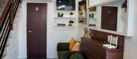 A modern entryway with a wooden door, a staircase, a green sofa with cushions, a small round plant, a mirror, and a mounted flat-screen TV.