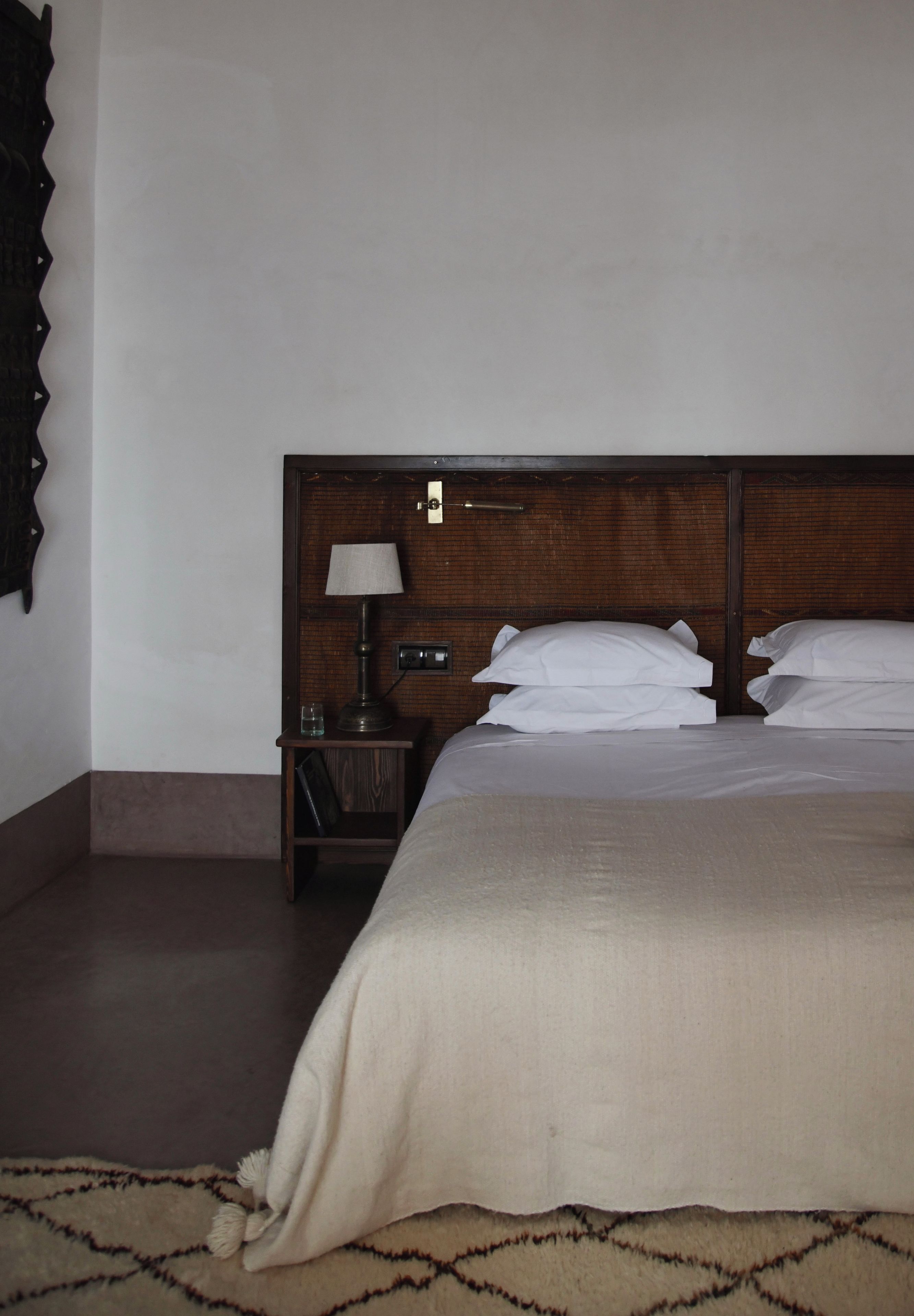 Habitación de hotel con cabecera de madera, una cama individual con ropa blanca, una mesita de noche con lámpara y una pequeña mesa con un vaso y un libro.