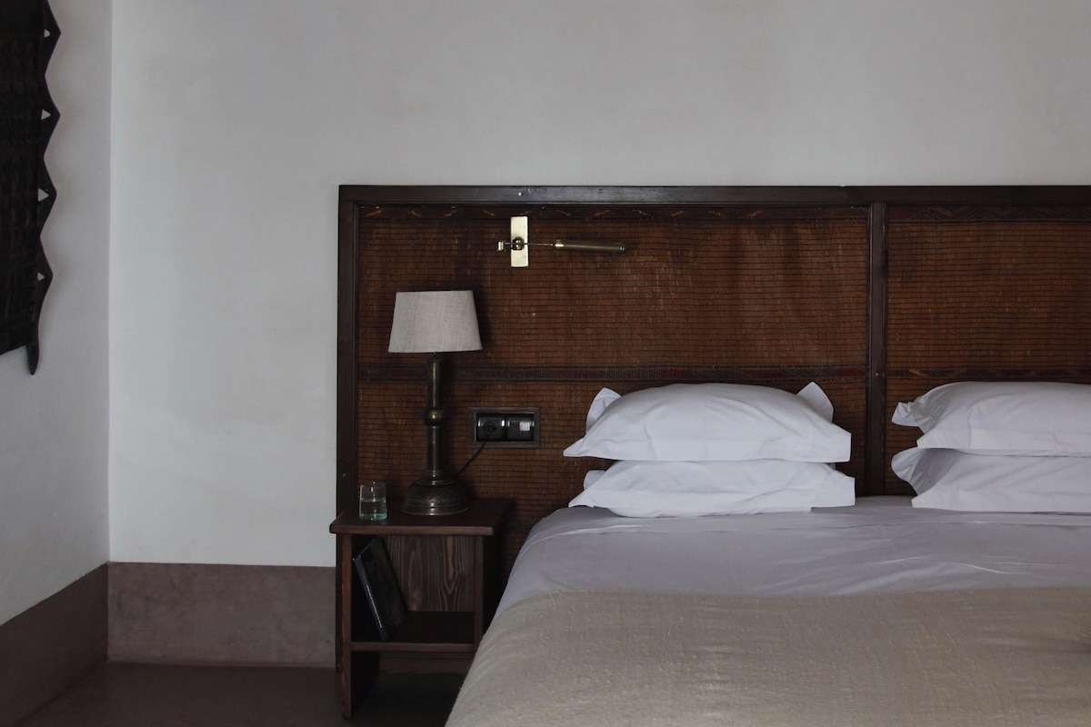 Een hotelkamer met een houten hoofdeinde, een enkel bed met witte lakens, een nachtkastje met een lamp en een klein tafeltje met een glas en een boek.