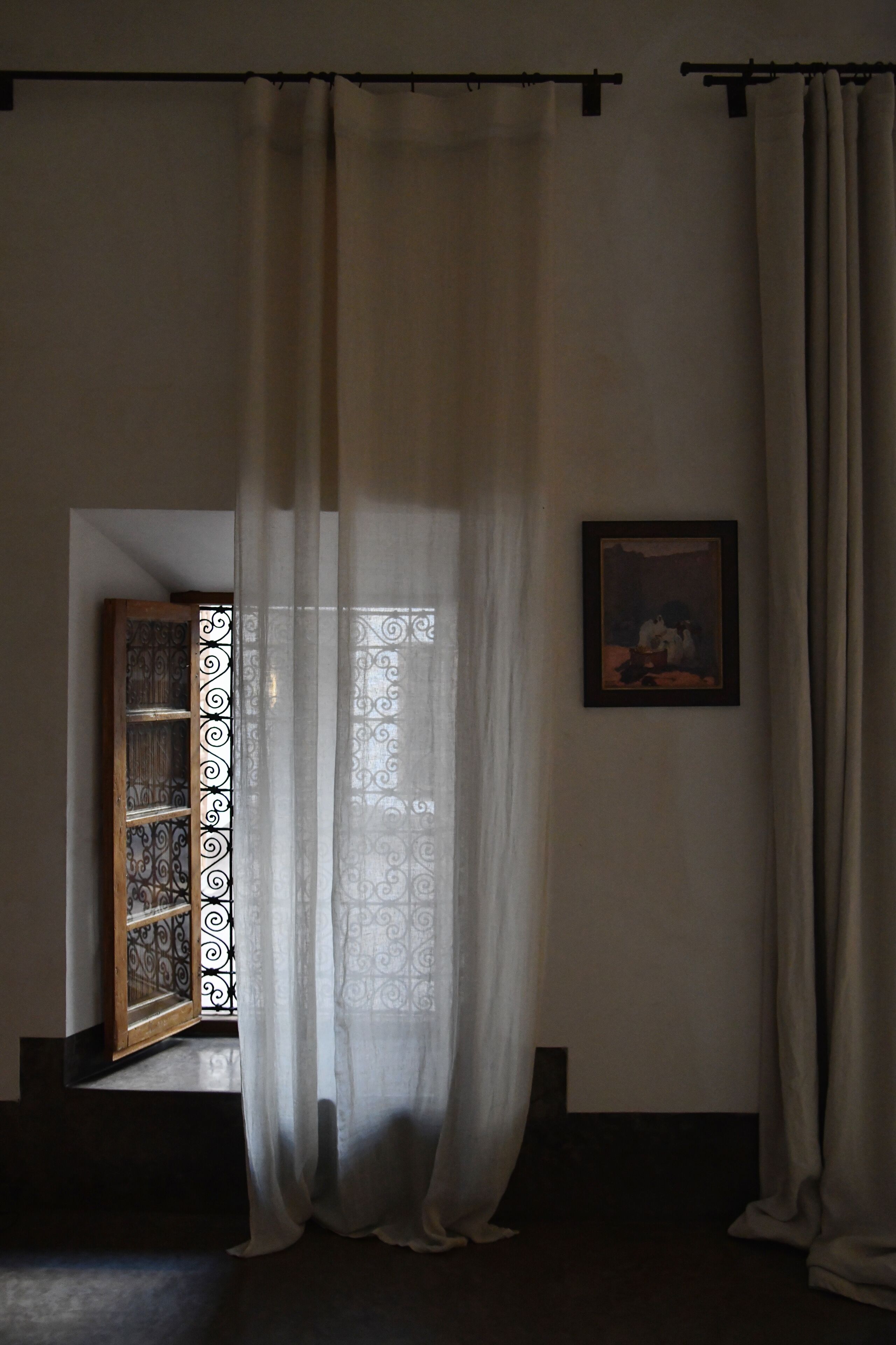Ein Zimmer mit einem großen Fenster, das mit durchsichtigen Vorhängen bedeckt ist, einem gerahmten Gemälde an der Wand und einem dekorativen Metallgitter.