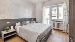 A modern bedroom with a large bed, a nightstand with a plant, a radiator, and a view of buildings through the window. - Rome Experience (Rome)