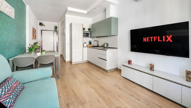 A modern living room with a teal sofa, a flat-screen TV, and a kitchen area with white cabinets and a microwave. - Rome Experience (Rome)