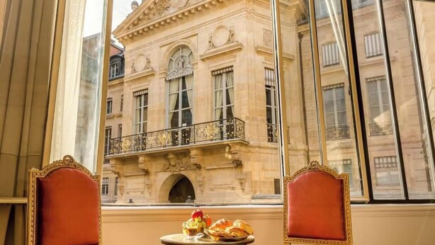 Una habitación con vistas a un edificio histórico, dos sillones tapizados en rojo y una pequeña mesa con un frutero y pasteles.