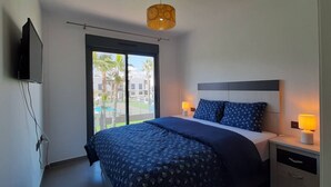 Ein modernes Schlafzimmer mit einem großen Bett, zwei Nachttischlampen, einem an der Wand montierten Fernseher und einem Fenster mit Blick auf Palmen und Gebäude.