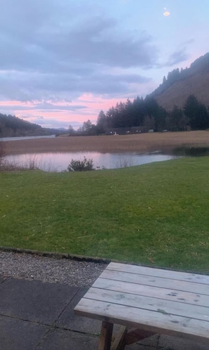 Une table de pique-nique se trouve dans une zone herbeuse, avec un lac et des collines en arrière-plan.