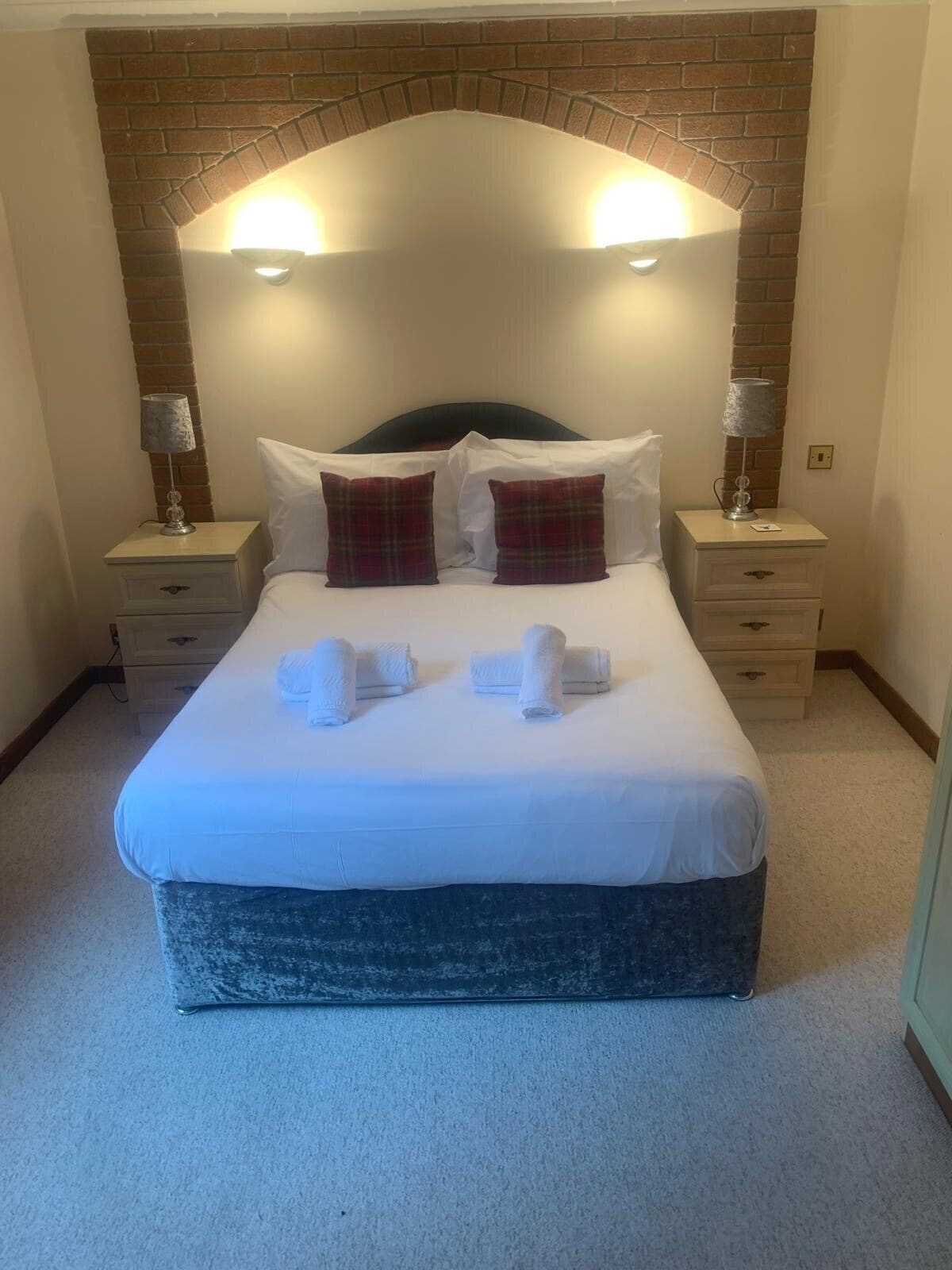 A neatly made bed with white linens and two red plaid pillows, flanked by two bedside tables with lamps, in a room with a brick-patterned headboard.