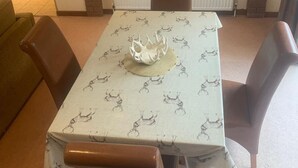 Et spiseområde med et bord dækket til fire personer, omgivet af stole. En dekorativ midtstykke står på bordet. Værelset har en murstenspejs med et indrammet billede over og en hvid radiator under.