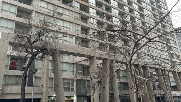 Un immeuble de grande hauteur doté de nombreux balcons, un magasin en rez-de-chaussée et des voitures garées le long de la rue.