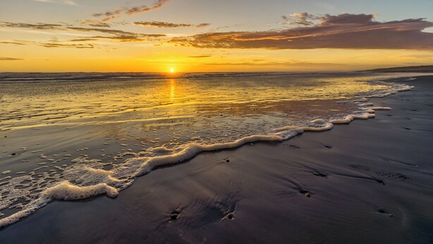 Ein Strand mit Sonnenuntergang, Wellen und Fußabdrücken im Sand.