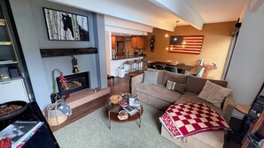 A living room with a fireplace, a sofa, a coffee table, and a dining area with a kitchen in the background.