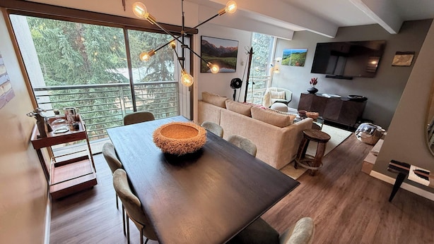 A modern dining area with a wooden table, chairs, and a decorative centerpiece. There is a sofa and a television in the background.