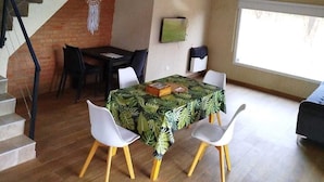 Un coin repas avec une table recouverte d’une nappe à motifs tropicaux, deux chaises blanches et un escalier menant à un niveau supérieur.