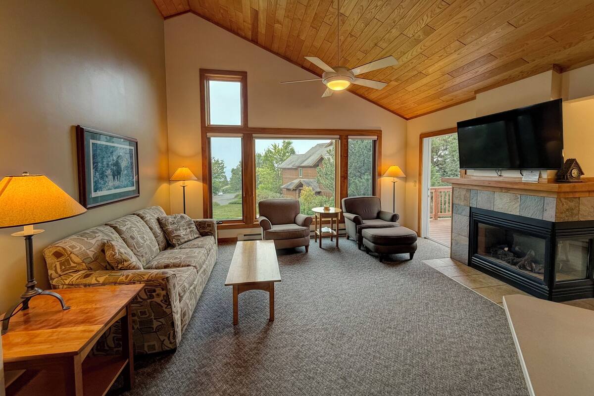 A cozy living room with a wooden ceiling, a fireplace, a television, and a large window offering a view of the outdoors.