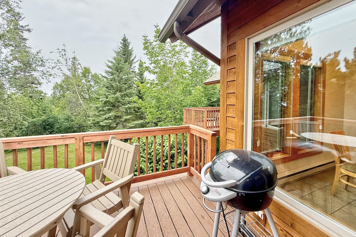 A wooden deck with a table and chairs, a grill, and a glass door leading to an indoor area.