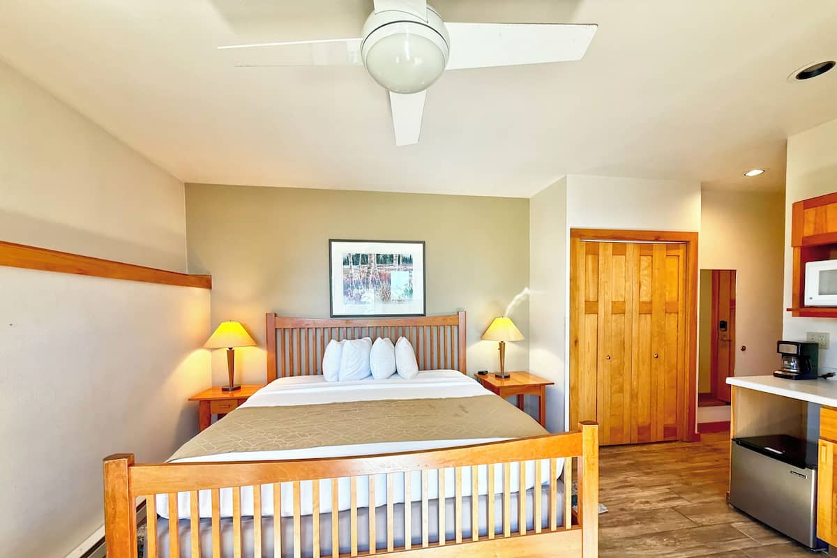 A hotel room with a bed, two bedside lamps, a ceiling fan, a framed picture on the wall, a wooden door, and a kitchenette area.
