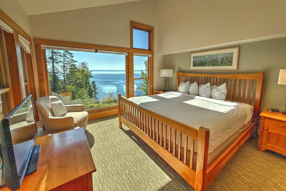 A bedroom with a large window offering a view of the ocean and trees, a wooden bed, a desk, and a chair.