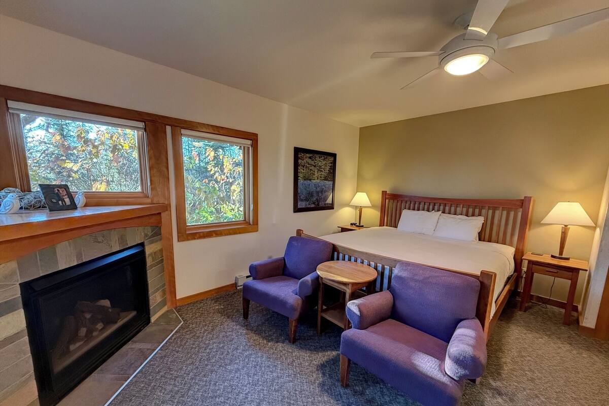 A hotel room with a large bed, two armchairs, a small table, a fireplace, and two windows with a view of trees.