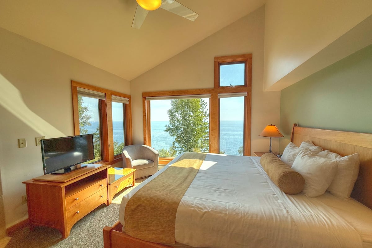 A bedroom with a large bed, a TV on a wooden stand, a chair, and a view of the sea through large windows.
