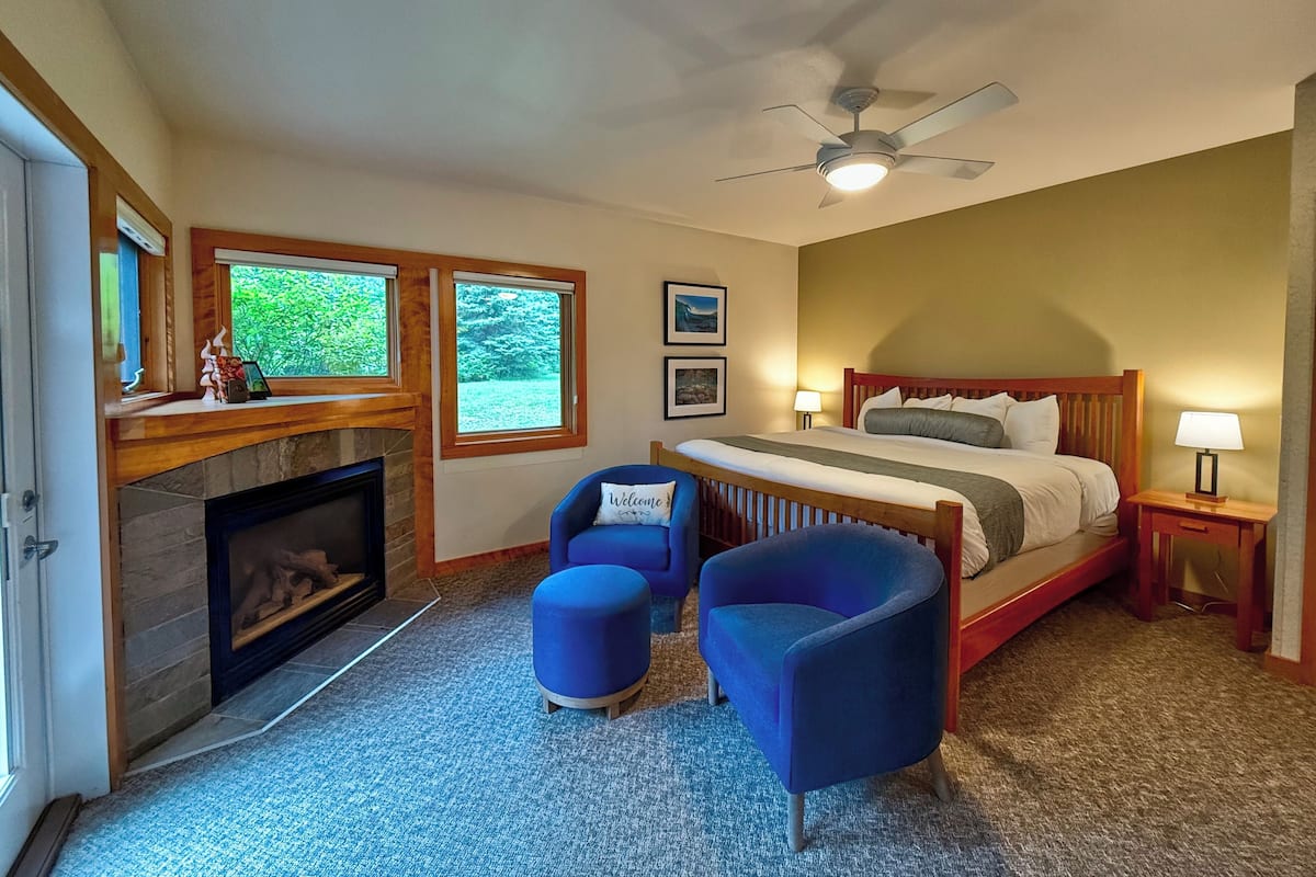 A bedroom with a fireplace, a bed, a ceiling fan, and two blue chairs.