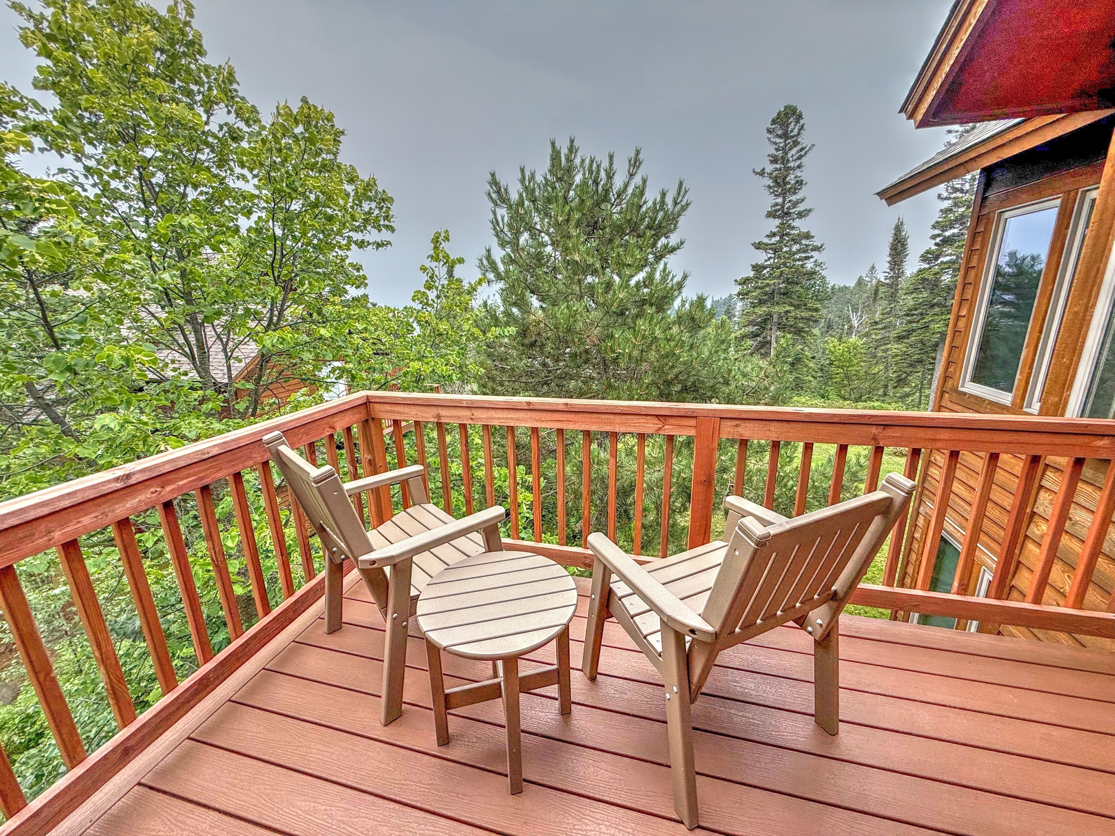 A wooden deck with a table and four chairs, surrounded by a railing, overlooking a forested area.
