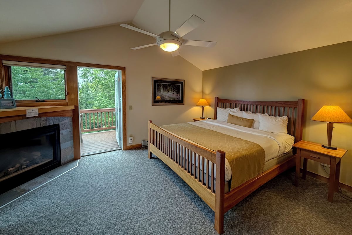A bedroom with a wooden bed, two bedside lamps, a ceiling fan, a fireplace, and a large window with a view of greenery.