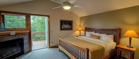 A bedroom with a wooden bed, two bedside lamps, a ceiling fan, a fireplace, and a large window with a view of greenery.