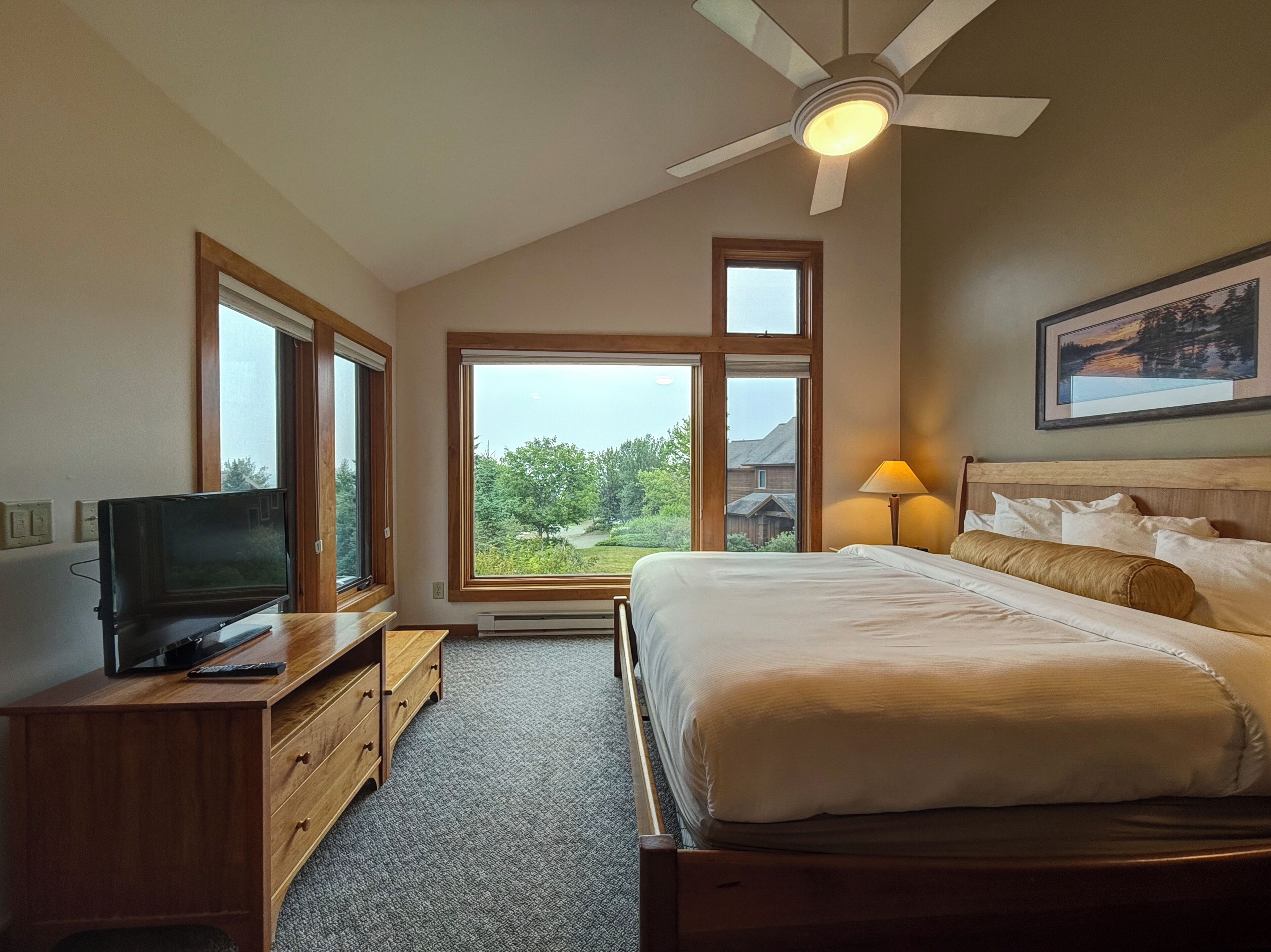 Un dormitorio con cama, televisor sobre una cómoda de madera, ventilador de techo y ventanas amplias con vistas a la vegetación y una casa.