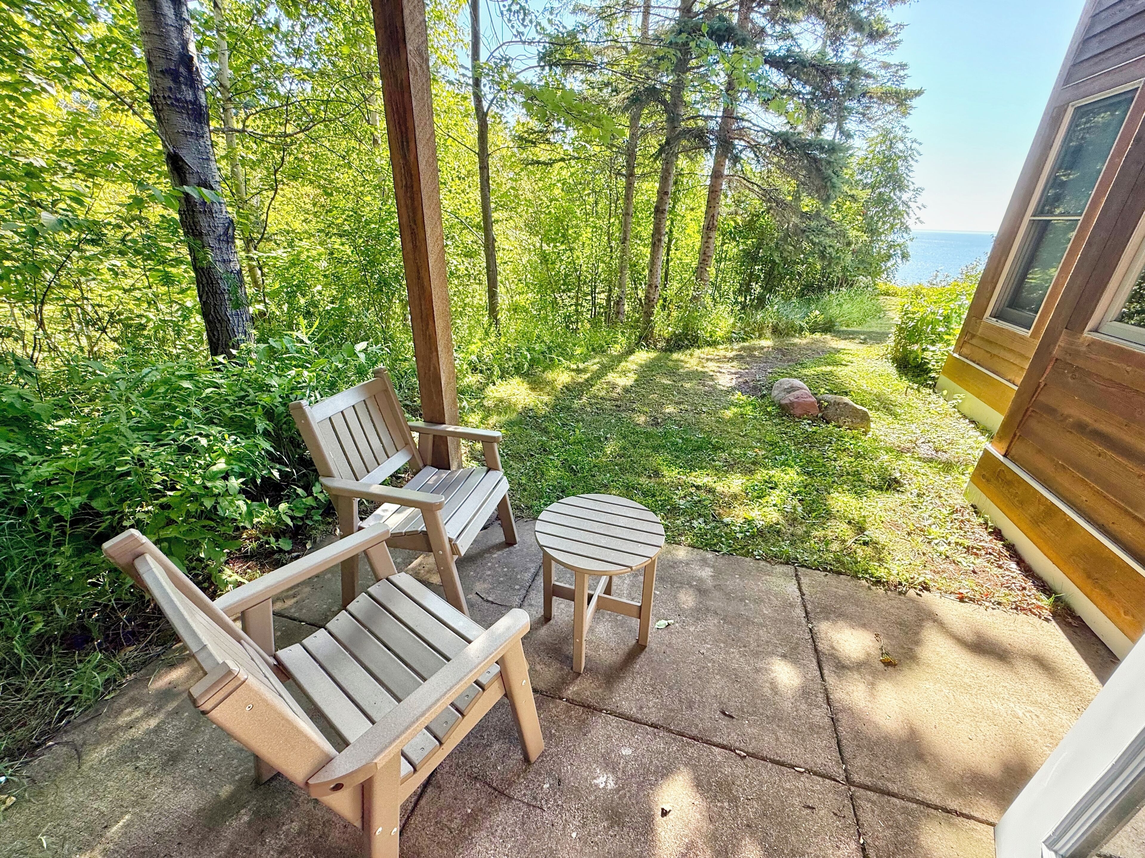 A wooden deck with two chairs and a small table, surrounded by lush greenery and a view of the ocean.