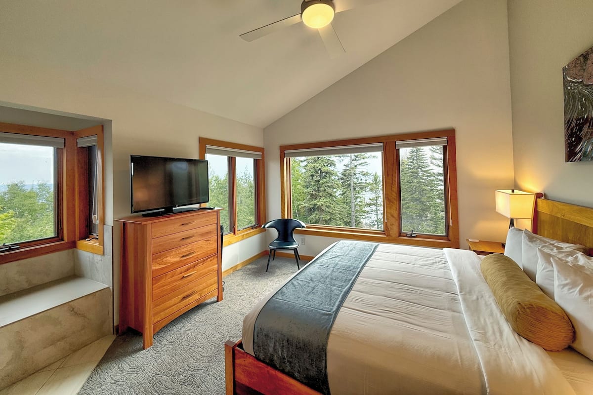 A bedroom with a bed, a TV on a wooden dresser, a chair, and large windows with a view of trees.