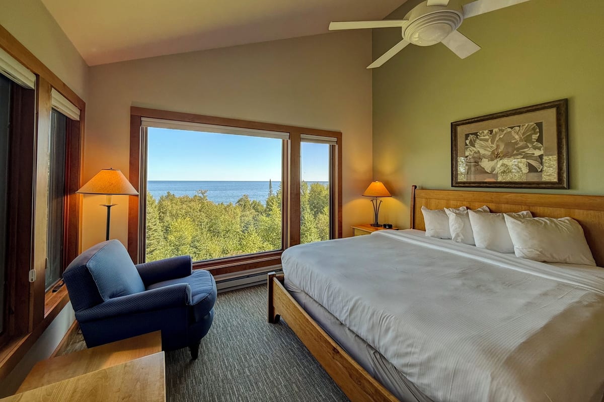 A hotel room with a large bed, two bedside lamps, a ceiling fan, a framed artwork, and a view of the ocean through large windows.