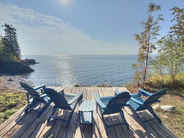 A wooden deck with two Adirondack chairs and a small table, overlooking a rocky shoreline and the sea. - Poplar River Condos (Lutsen)