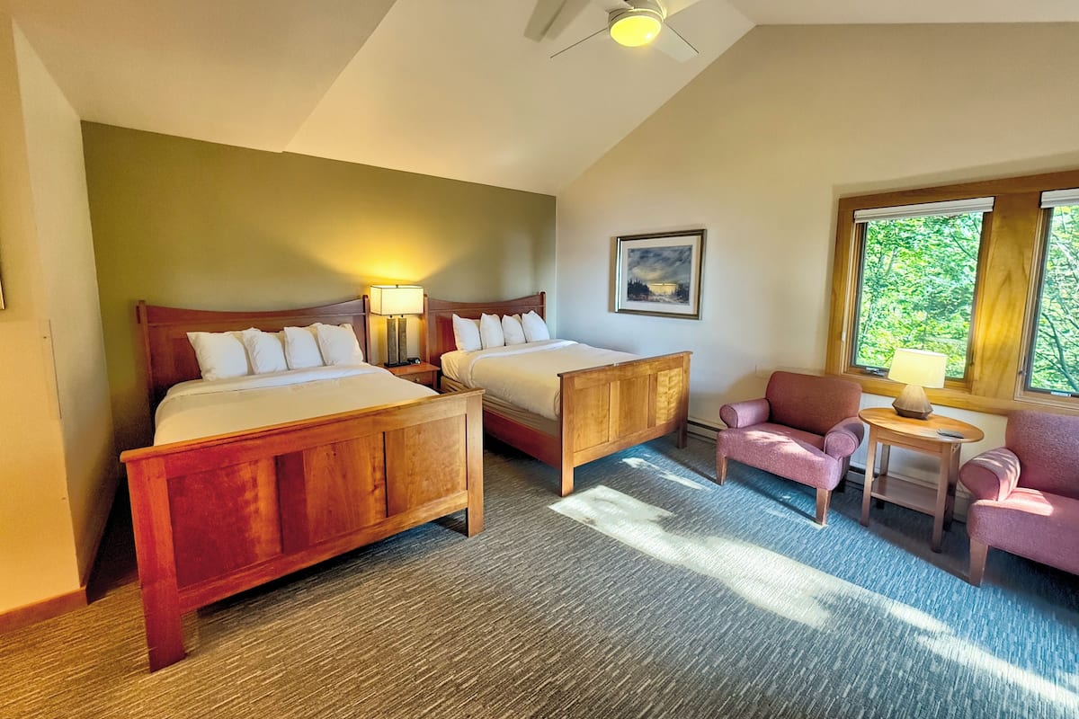 A hotel room with two wooden beds, a small desk with a lamp, a chair, and a window with a view of trees.