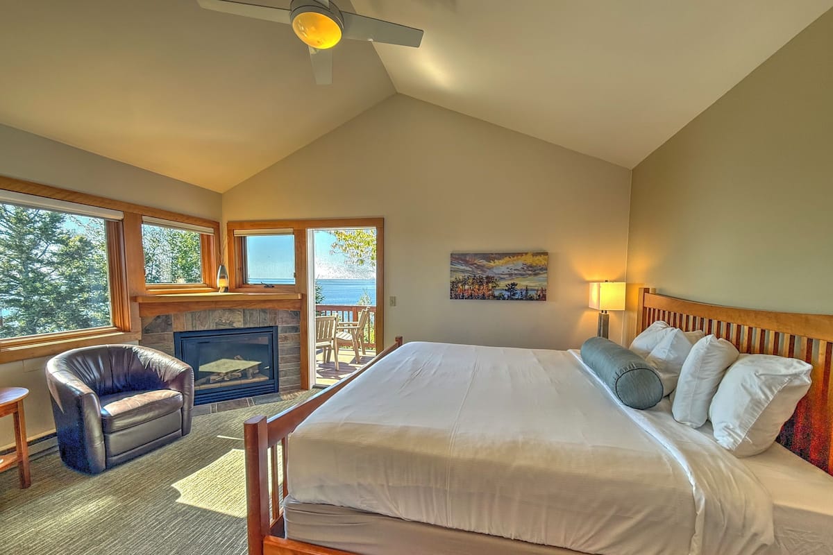 A bedroom with a large bed, a fireplace, a chair, and a view of the outdoors.