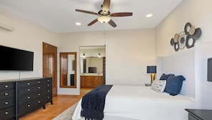 Une chambre à coucher avec un lit, une télévision, un ventilateur de plafond, une commode et une salle de bain visible à travers une porte ouverte.
