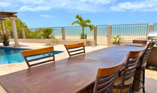An outdoor dining area with a pool, chairs, and a view of the beach.