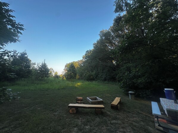 An outdoor seating area with picnic tables and benches, surrounded by trees and grass.