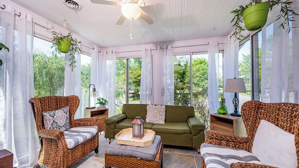 Ein Wintergarten mit Korbmöbeln, Deckenventilator und großen Fenstern mit Blick auf Grünflächen.