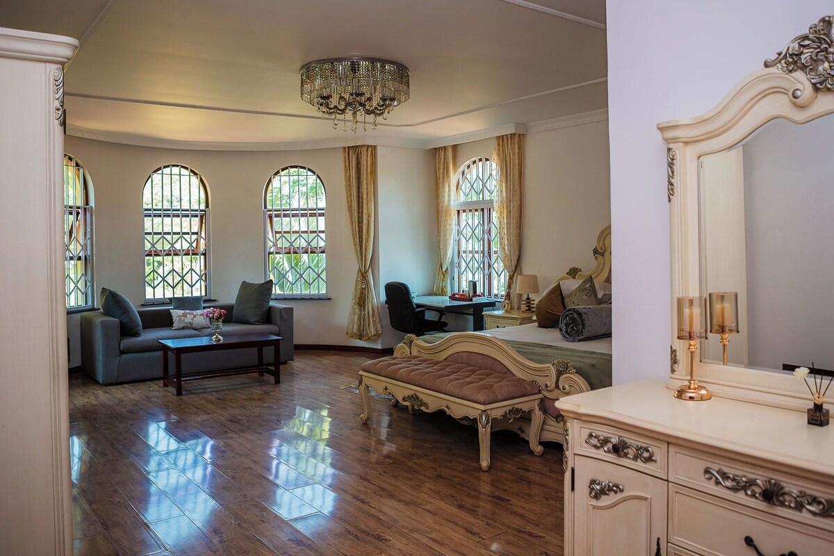 A spacious room with a wooden floor, a large bed with ornate headboard, a desk with a chair, and a sofa.