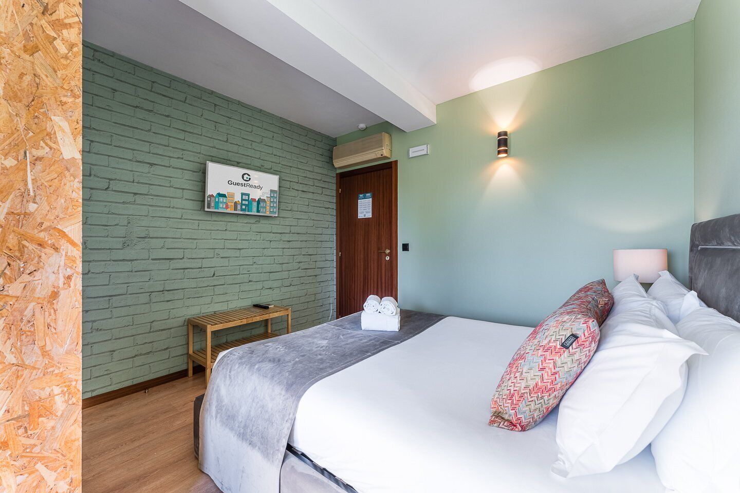 A hotel room with a bed, a wooden nightstand, a green brick wall, and a sign indicating the room is ready for guests.