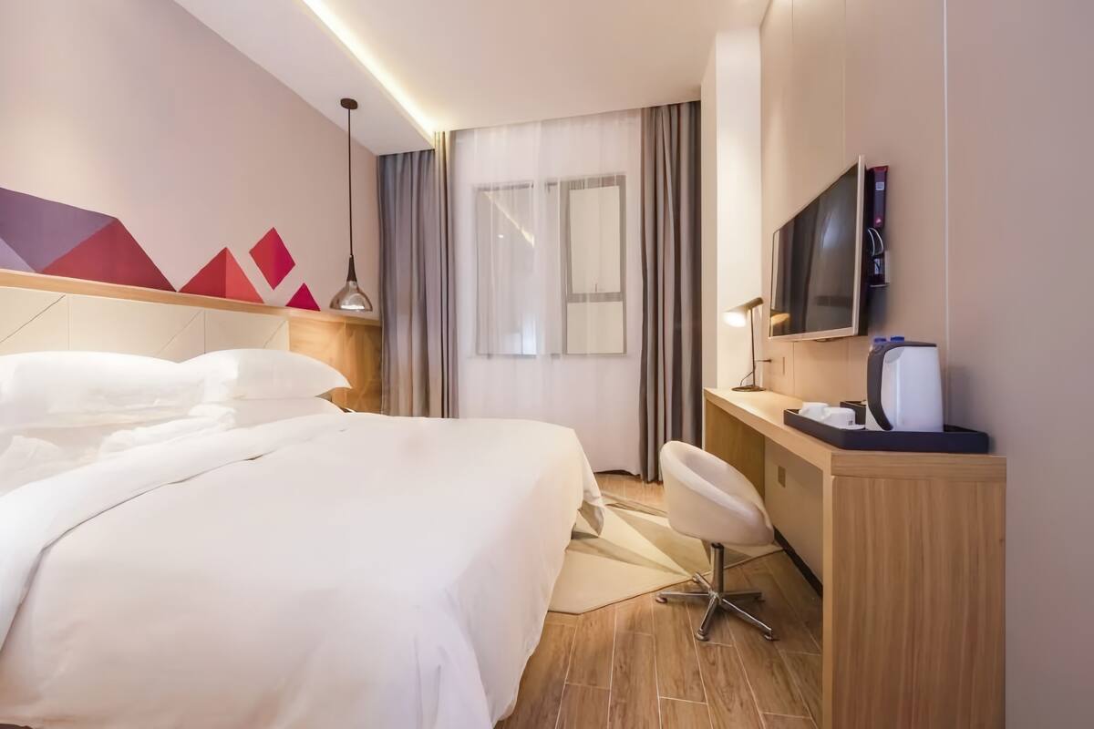 A modern hotel room with a large bed, a desk, a chair, a television, and a window with curtains.