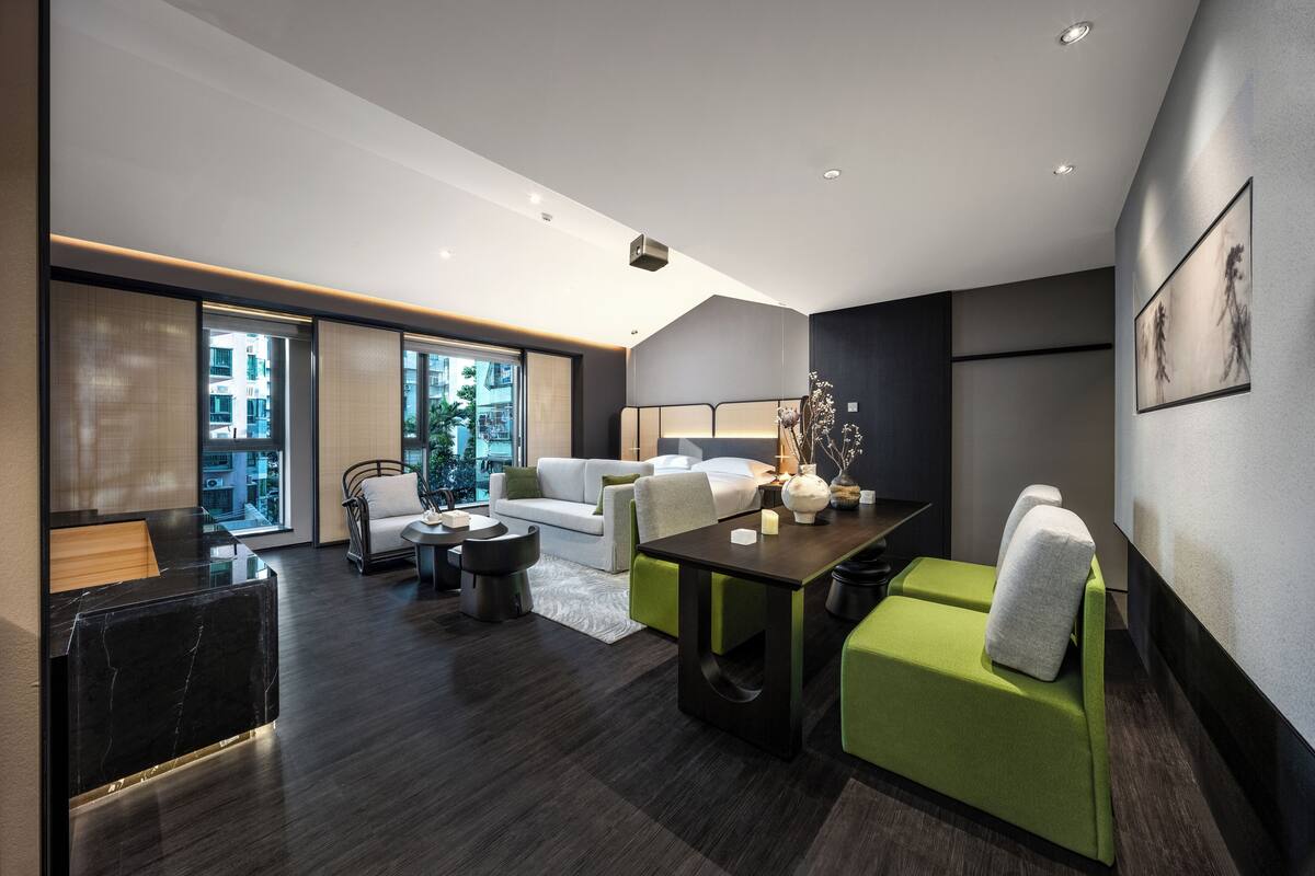 A modern hotel room with a dark wood floor, a green sofa, a white sofa, a black reception desk, and a large window offering a city view.