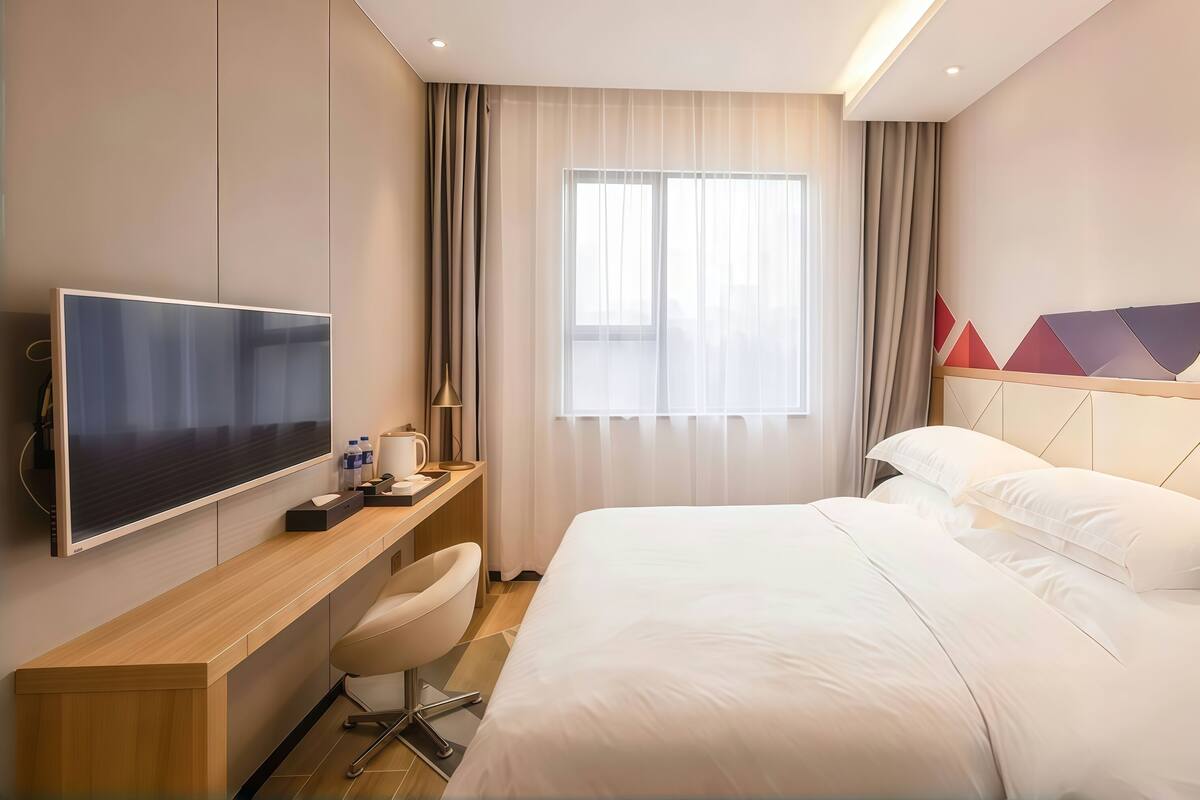 A modern hotel room with a large bed, a flat-screen TV mounted on the wall, a desk with a chair, and a window with sheer curtains.