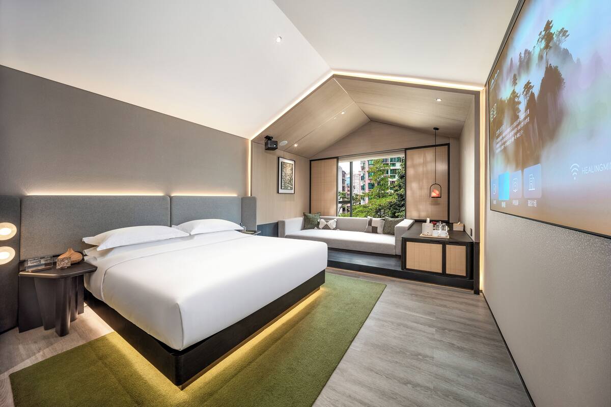 A modern hotel room with a large bed, a sofa, a desk, and a large window.